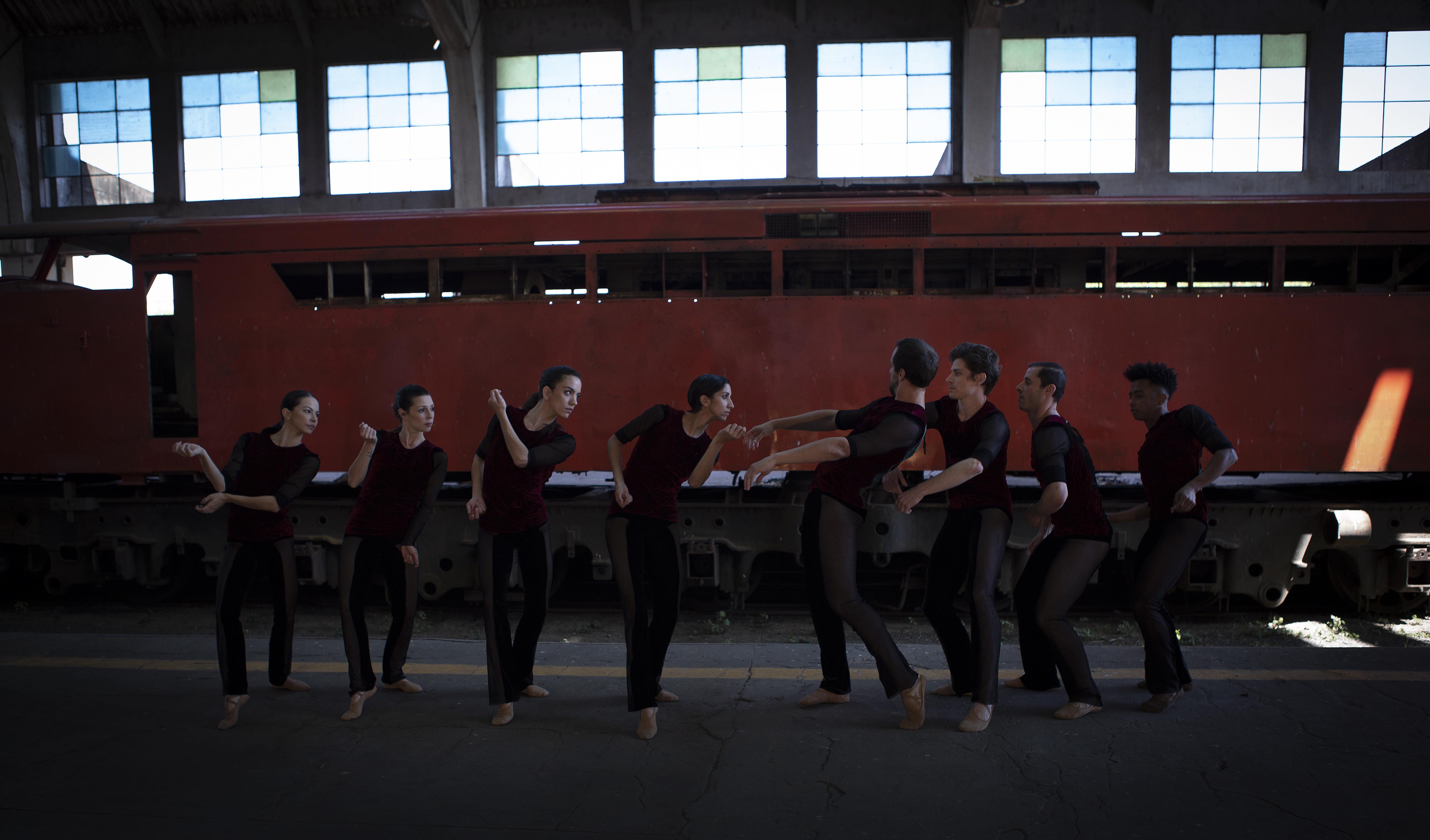 Em frente à lateral de um trem vermelho, quatro bailarinas e quatro bailarinos estão dispostos em fila indiana com calças pretas e blusas vermelhas