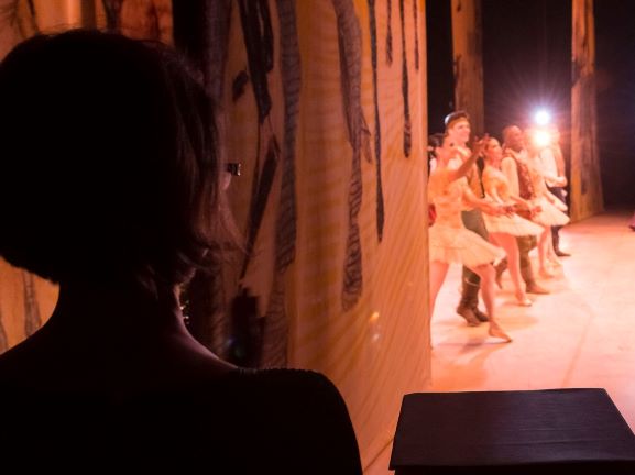 À esquerda, silhueta de Inês Bogéa, de cabelos curtos, olhando de dentro da coxia para o palco iluminado e com a presença dos bailarinos Foto Wilian Aguiar